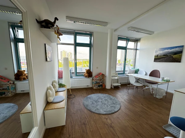 Ansicht im Therapieraum 1, weiße Wände, brauner Boden, zwei fast raumhohe Fenster, Schreibtisch, Stühle und Bilder