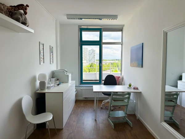 Ansicht im Therapieraum 2, weiße Wände, brauner Boden, zwei fast raumhohe Fenster, Schreibtisch, Stühle und Bilder